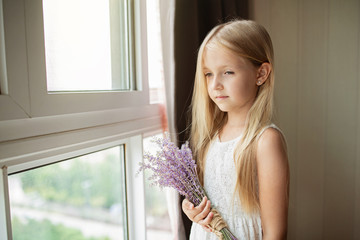 Cute little caucasian girl with blonde hair holding bouquet of purple flowers at home. Kid standing near window during coronavirus covid-19 pandemic quarantine.