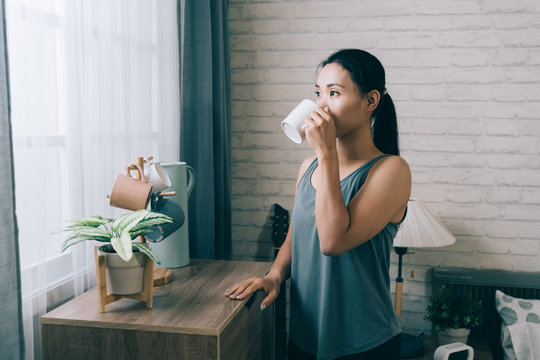 asian sportive woman standing by window placing hand on cupboard is having drink after workout. chinese female coach looking outside is taking a break with a cup of boiled water. - Powered by Adobe
