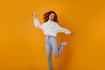 Fototapeta na wymiar Gorgeous girl in blue jeans jumping in studio. Indoor photo of dreamy ginger woman dancing on orange background.