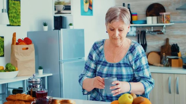 Grandmother taking cup of hot coffee for counter in kitchen during breakfast and looking at camera with a smile. Authentic portrait of relaxed elderly older person in the morning, enjoying fresh warm