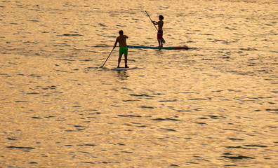 Recreation on the sea.  During hot summer weather two people maneuver their paddle boards across the golden hour water as the sun begins to set.