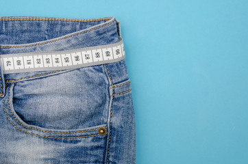 Blue jeans with a white measuring tape instead of a belt on a blue background, copy space, Top view. Concept of a healthy lifestyle and diet.