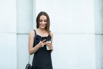 Photo of smiling young woman wearing sunglasses walking outdoors with coffee chatting by mobile phone.