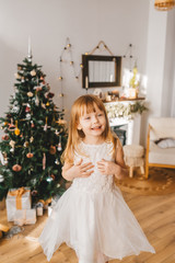 Beautiful little girl with long hair standing near the Christmas tree, in a beautiful white dress, cute smiles.