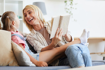 Family of two reading a book together and laughing while resting on sofa at home