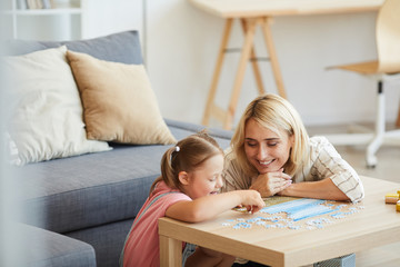 Young happy mother teaching her daughter with down syndrome collecting puzzles at the table in the...