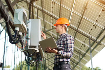 Expert is inspecting quality of solar panels. Man in hardhat standing with laptop near electrical equipment. Solar station sunny power electricity. Innovation future.