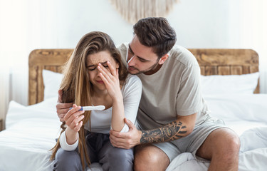 Sad man hugs crying woman and looks at pregnancy test in bedroom interior - obrazy, fototapety, plakaty