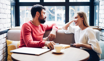 Caucasian male and female sitting at cafeteria table and discussing trip plan during leisure time in cafe interior, positive boyfriend and girlfriend spending day in public coffee shop with wifi