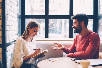 Young woman writing checklist of stuff while planning trip with boyfriend during dating in coffee shop, best friends discussing ideas for event noting in dairy while sitting together in cafe interior