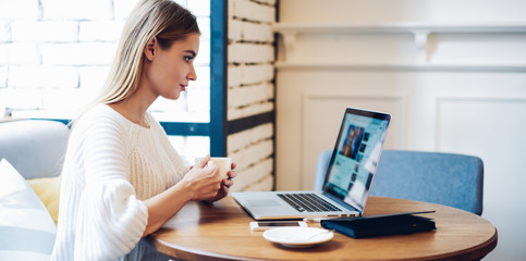 Side view of millennial hipster girl dressed in casual apparel watching webinar video during leisure time in public coffee shop with wifi internet for users, female generation reading news on website