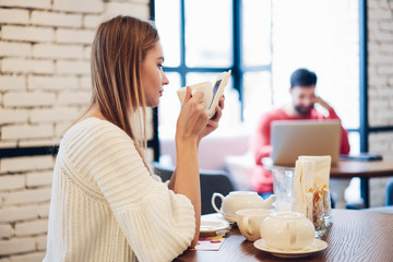 Side view of attractive female holding cup with hot tasty tea and writing email message via cellphone application connecting to wifi internet in coffee shop, millennial hipster girl checking media