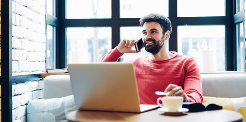 Positive male freelancer making cellphone call sitting with modern laptop computer in coworking...