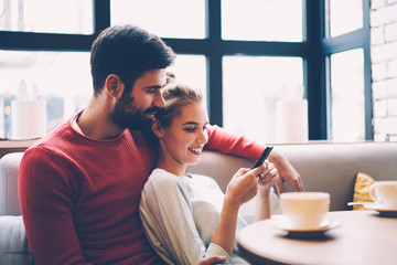 Smiling hipster girl showing web page of store with item for buying to his boyfriend spending time together, couple in love booking tickets for movie via smartphone application resting in cafe