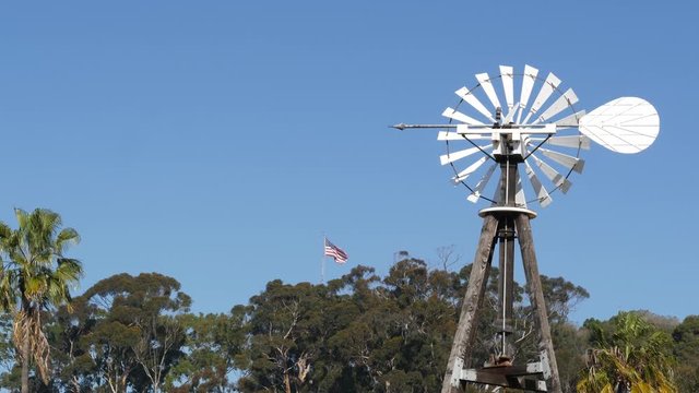 Classic retro windmill, bladed rotor and USA flag against blue sky. Vintage water pump wind turbine, power generator on livestock ranch or agricultural farm. Rural symbol of wild west, rustic suburb.