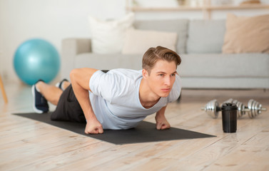 Concentrated guy push up on fists on mat in interiowith dumbbells, ball and bottle of water