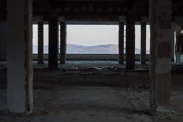 A view of the mountains on the horizon with a blue sky through a large window at the end of a row of columns on an abandoned building on the outskirts of the city.
