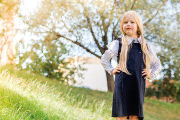 A little blonde girl with two ponytails, in a school uniform, posing against the backdrop of greenery, trees, sunny weather.