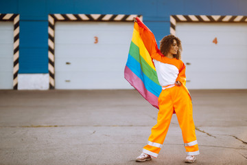 Young African American lesbian woman with LGBT rainbow flag in the street at sunset. Stylish woman...