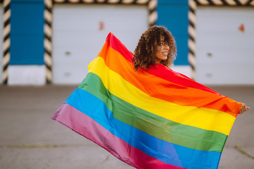 Young African American lesbian woman with LGBT rainbow flag in the street at sunset. Stylish woman with curly hair in an orange suit posing with lgbt pride flag.