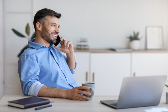 Smiling young businessman having coffee break at workplace and talking on cellphone