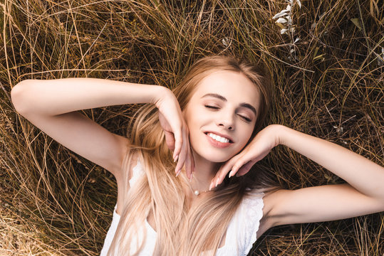 top view of sensual blonde woman touching face while lying on green grass with closed eyes