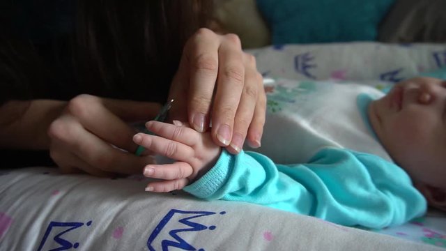 A mother cuts the nails of a newborn child. The woman carefully cuts her nails with baby scissors. The process takes place at home on the bed, the child lies in a cocoon. Sometimes she cries.