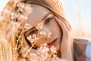 portrait of pensive blonde woman in straw hat touching face while looking at camera near wildflowers - Powered by Adobe