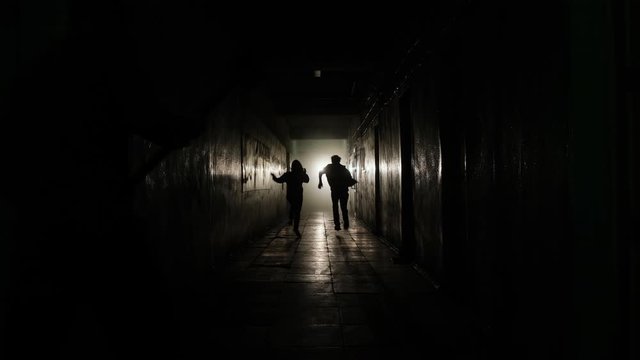 A general shot of a man and a girl running away along a straight corridor from the shadows into the light from a limping man with an ax. High quality 4k footage