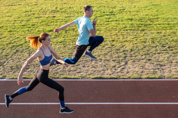 Obraz premium Young couple of fit sportsmen boy and girl running while doing exercise on red tracks of public stadium outdoors.