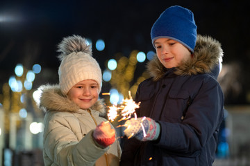 Two cute young children, boy and girl in warm winter clothing holding burning sparkler fireworks on...