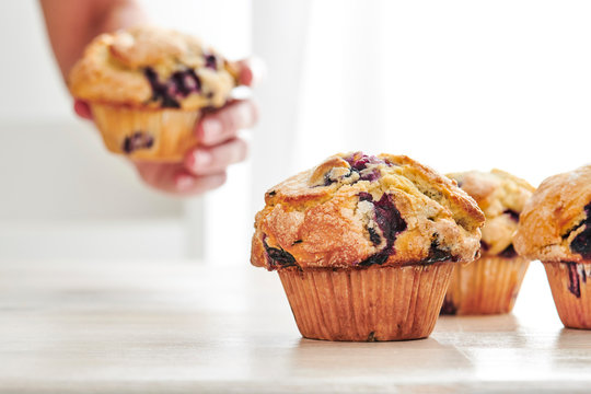 Rustic Blueberry Muffins On A White, Wood Table With Bright Window Light In The Background. A Woman’s Hand Grabs A Muffin Off The Table In The Background.