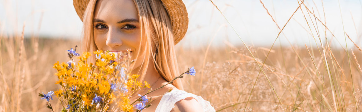 young blonde woman in straw hat looking at camera while holding wildflowers, horizontal image