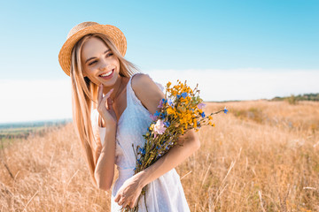 blonde, sensual woman in straw hat and white dress holding wildflowers and looking away in meadow