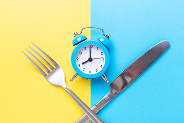 Blue alarm clock, fork, knife on colored paper background. Intermittent fasting concept