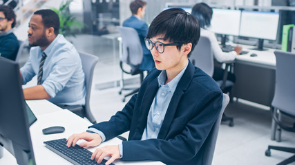 Young Professional Japanese Stock Broker Work on His Desktop Computer. Diverse and Motivated Business People Work in Modern Open Office.