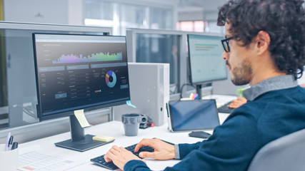 Young Handsome Manager with Curly Hair Works on a Desktop Computer in Accountant Software. Diverse...