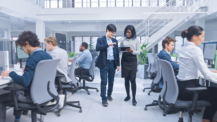 Couple of Young Male and Female Specialiasts Walk in the Office Holding a Tablet Computer, Ignore Their Colleague and Discuss Work. Motivated Diverse Business Managers in Modern Open Office. 