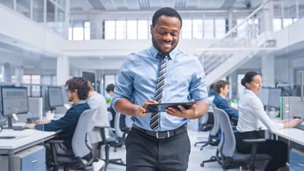 Handsome Young Black Manager in a Shirt Walking Pass His Business Colleagues with a Tablet and Supervise Their Work. Diverse and Motivated Business People Work on Computers in Modern Open Office.