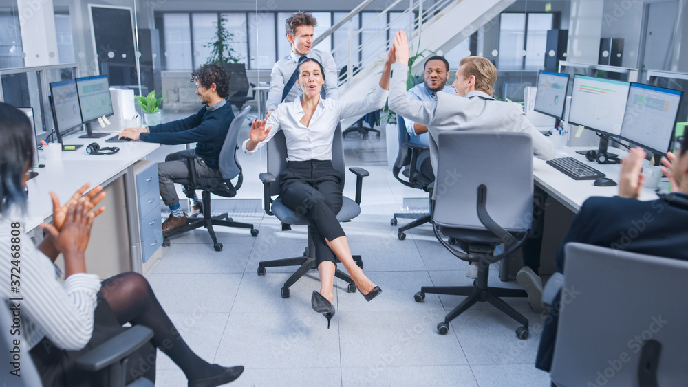 Poster cheerful employee pushes his beautiful female colleague on a chair between rows of desks and give a 