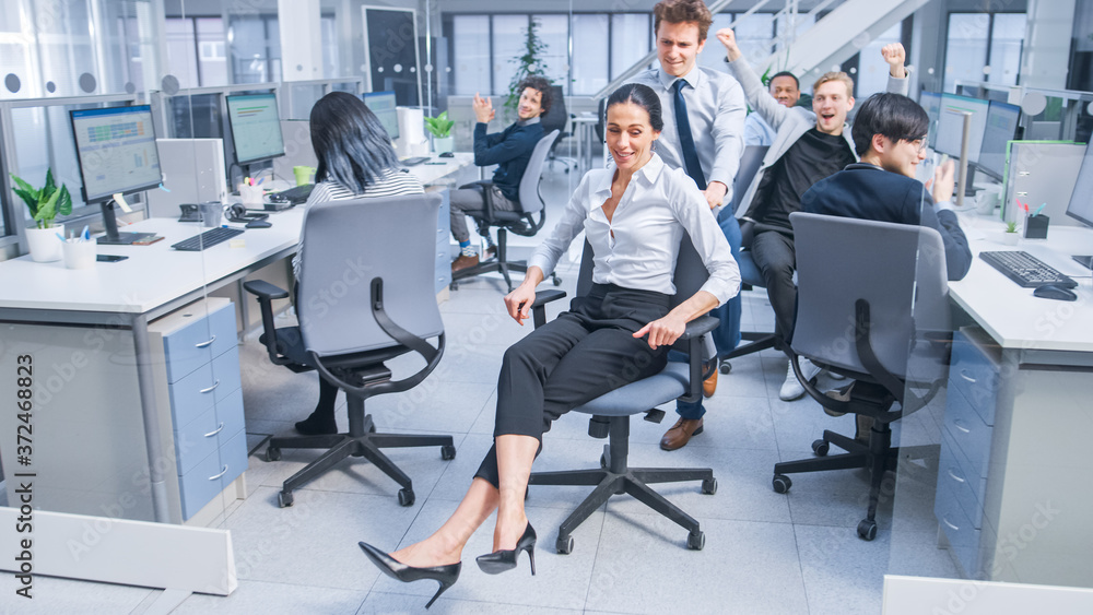 Poster cheerful employee pushes his beautiful female colleague on a chair between rows of desks with divers