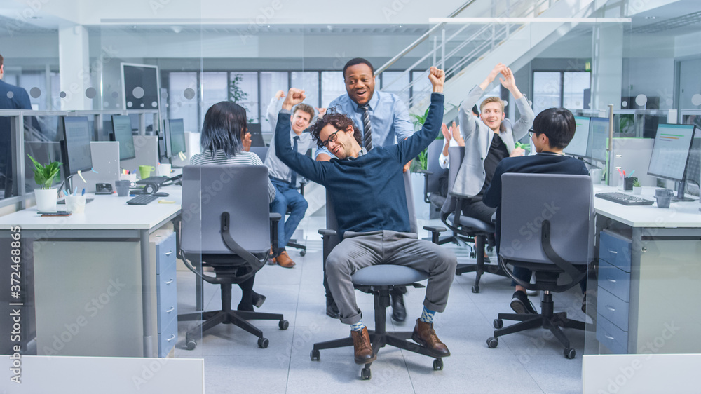 Poster Cheerful Employee Pushes His Handsome Colleague on a Chair Between Rows of Desks with Diverse Business People Working on Desktop Computers in Modern Office Space. 
