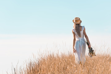 back view of woman in white dress and straw hat holding wildflowers while standing on grassy hill