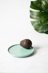 One whole avocado on a green plate against a background of tropical leaves. Light colors, minimalistic trendy composition