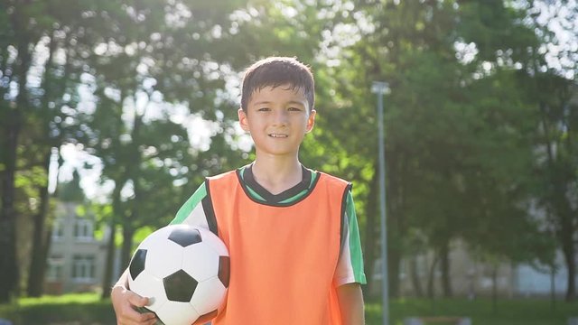 Front view of confident sporty teen boy in soccer sportswear which posing on camera with ball during football training on the stadium in the urban park