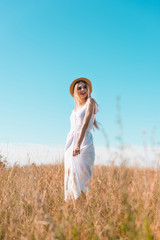Naklejka na ściany i meble selective focus of blonde woman in white dress and straw hat standing in field against blue sky and looking away