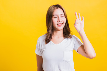 Asian Thai happy portrait beautiful cute young woman standing wear t-shirt showing gesturing ok sign with fingers looking to away, isolated studio shot on yellow background with copy space