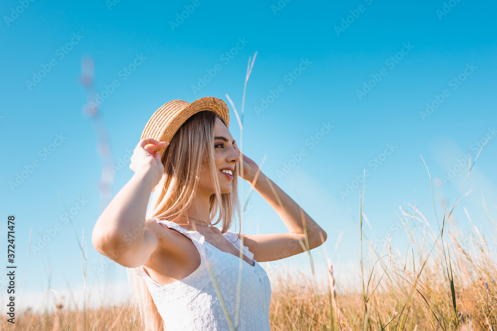 Wall mural selective focus of stylish blonde woman touching straw hat while looking away against blue sky