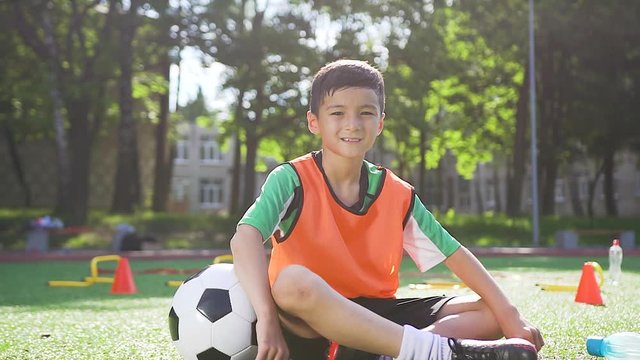 Likable contented asian teen boy in orange vest sitting on the artificail turf with football ball and looking at camera