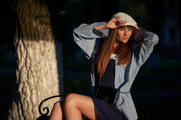 A young woman straightens her cap in the evening park while sitting on a bench.